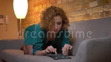 姜黄色卷发的白种人女孩躺在沙发上，在舒适的家专心地看着平板电脑的画像